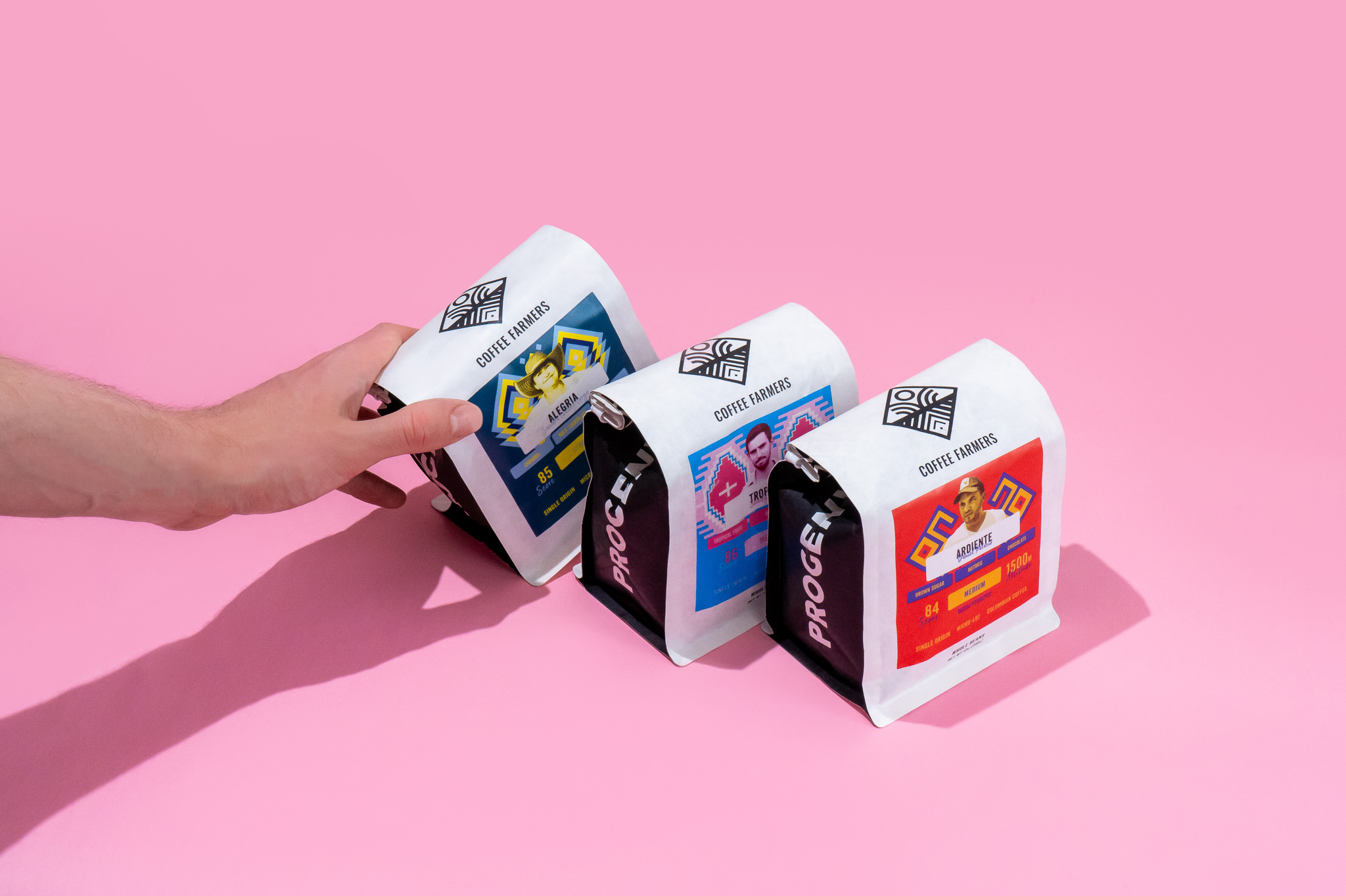 Three bags of Progeny Coffee on a pink backdrop. A hand is grabbing the bag on the left.