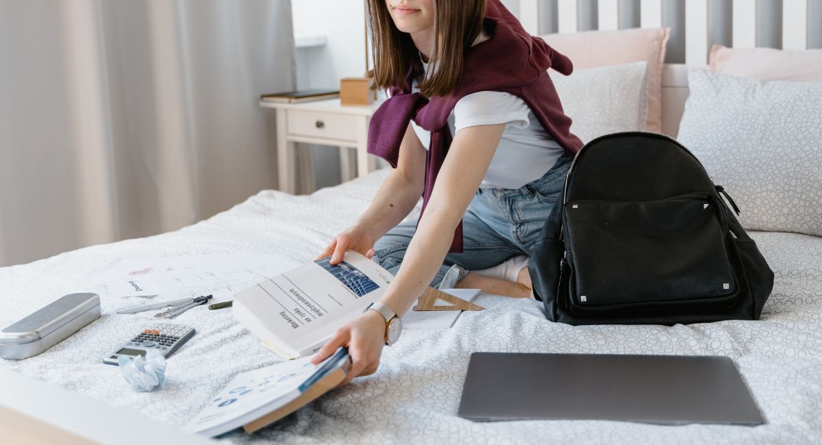 Woman sitting on a bed with her textbooks, laptop, and backpack.