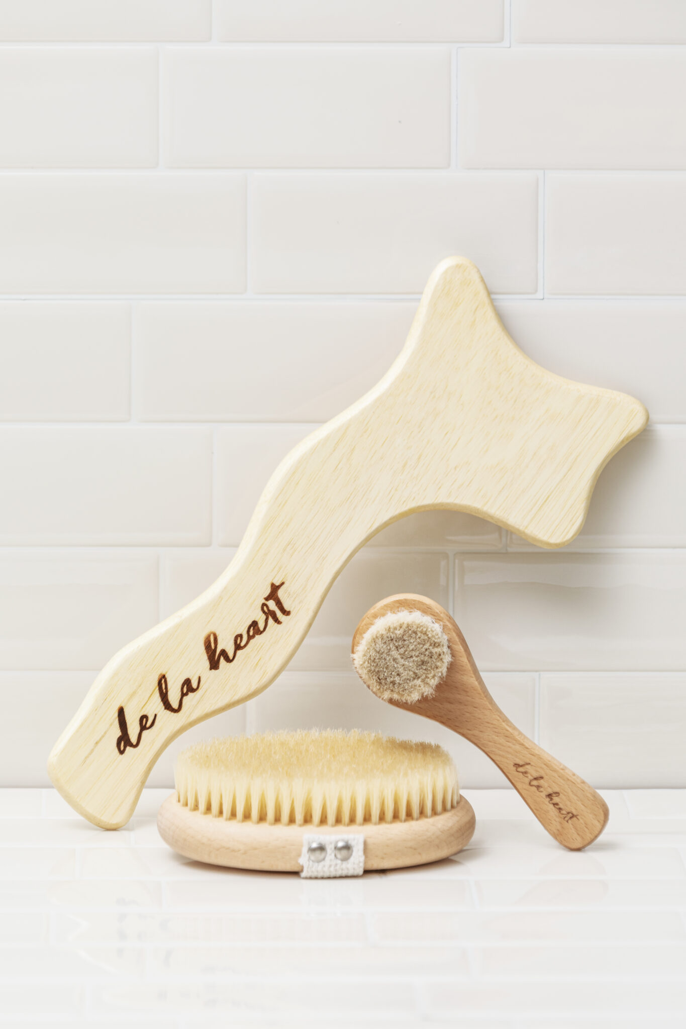 De La Heart's Body Massage Tool, body and facial dry brushes leaning against a white tile background.