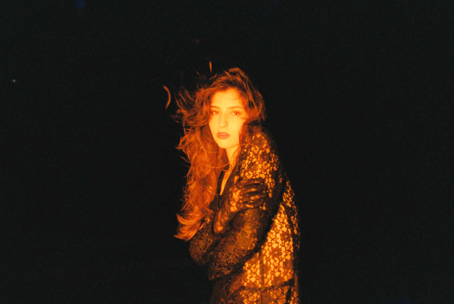 Artful promotional image of Andrea Bejar in a dark setting with a black background. Lighting is warm, her hair is blowing, and she is wearing a black outfit with a crocheted floral pattern.