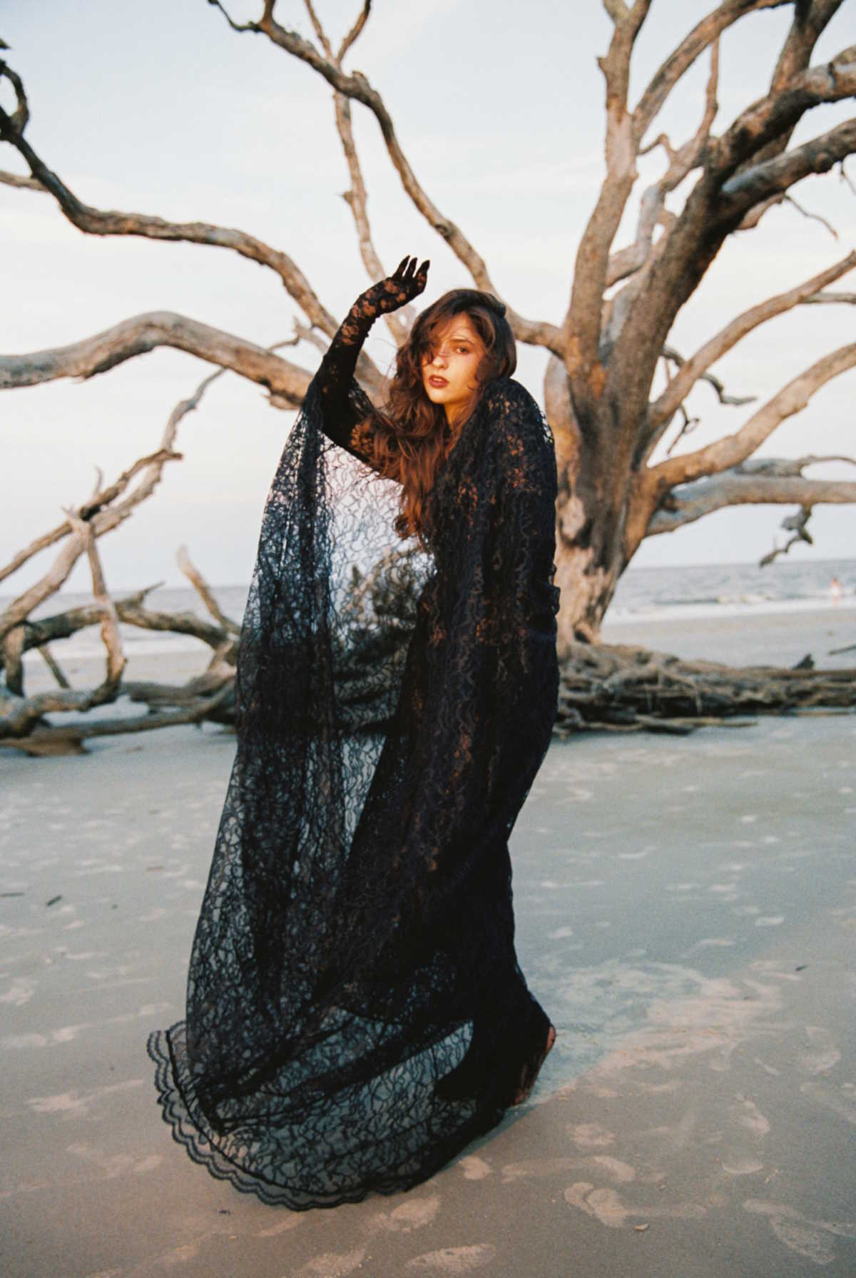 Artful promotional image of Andrea Bejar standing on a beach. The weather appears to be overcast, and a large tree that appears to be dead is in the background. Andrea is wearing a long, flowy black crocheted dress.