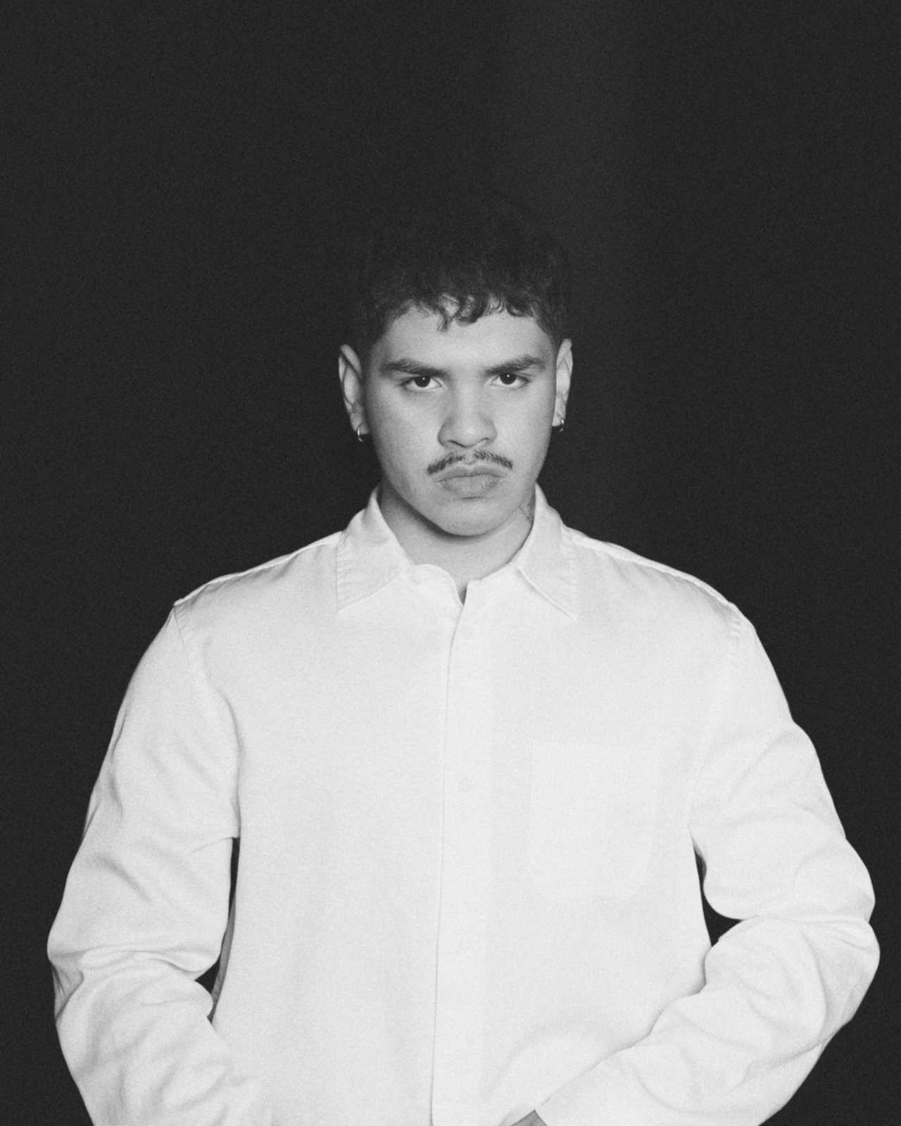 Promotional image of Andry Kiddos. The background is black. The image is black and white. He is standing and wearing a long-sleeve white button-down shirt.