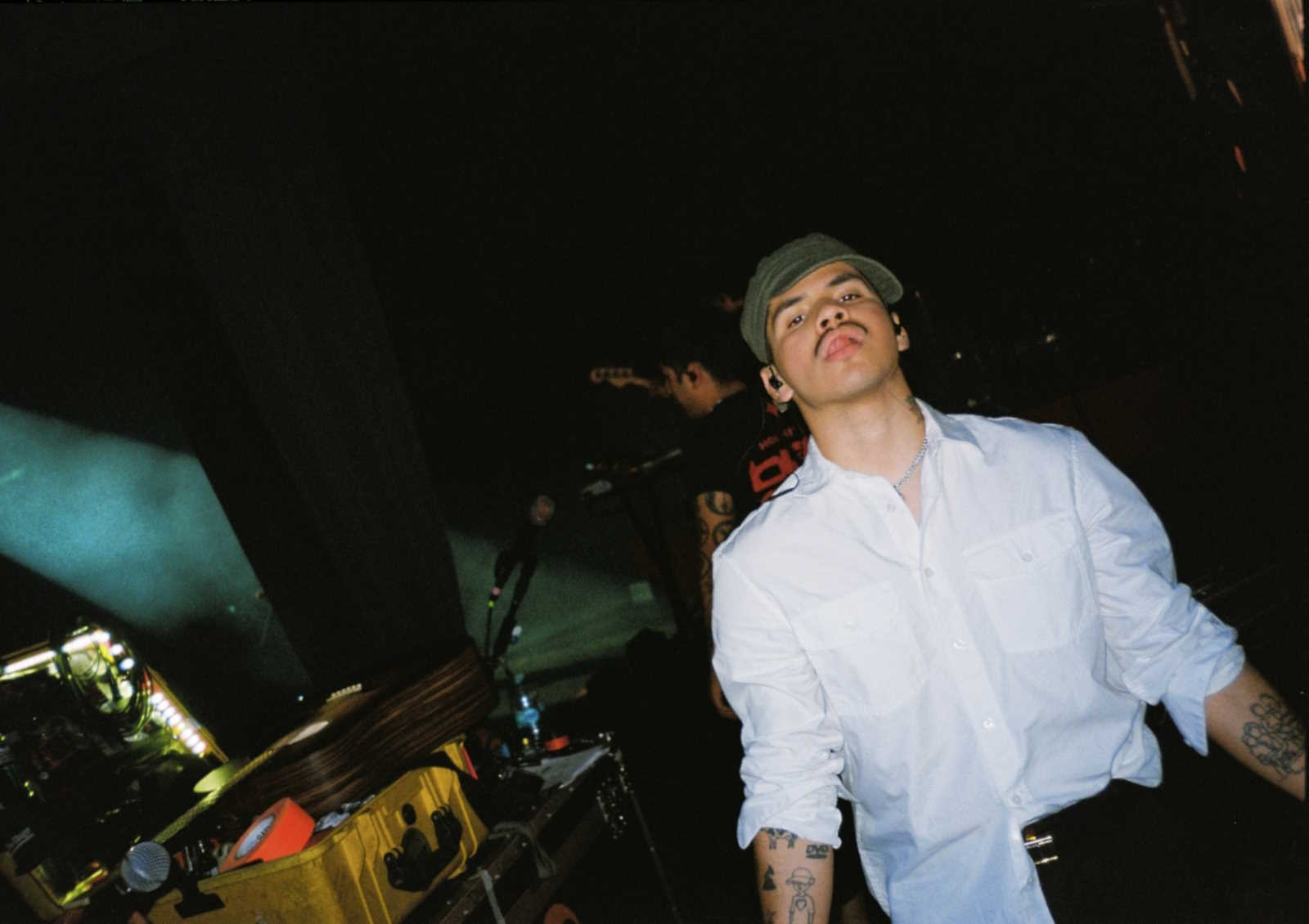 Promotional image of Andry Kiddos. The environment is dark, maybe a studio or nightclub. He is standing in front of what appears to be music or DJ equipment.