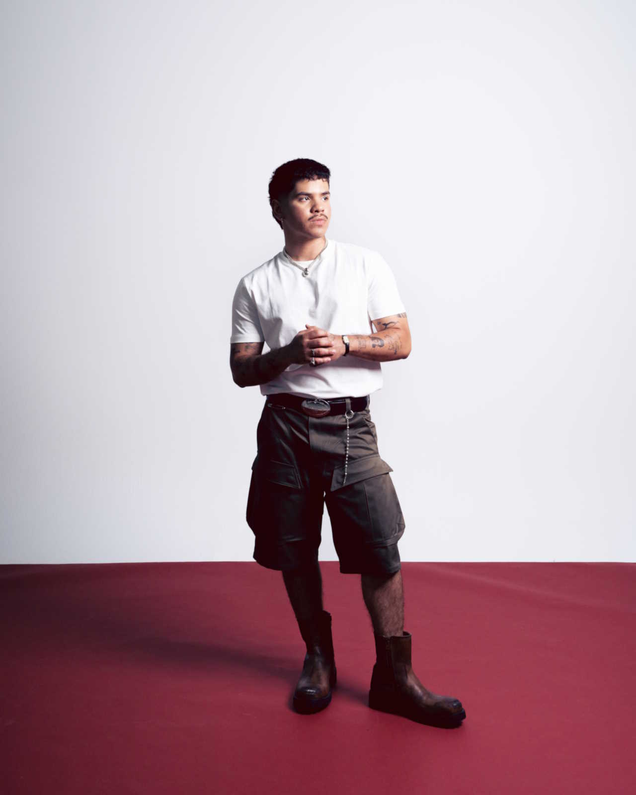 Promotional image of Andry Kiddos. He is stanging in front of a white wall and on a dark red floor that appears to be covered in a smooth cloth. He is wearing a white t-shirt and olive green cargo shorts.