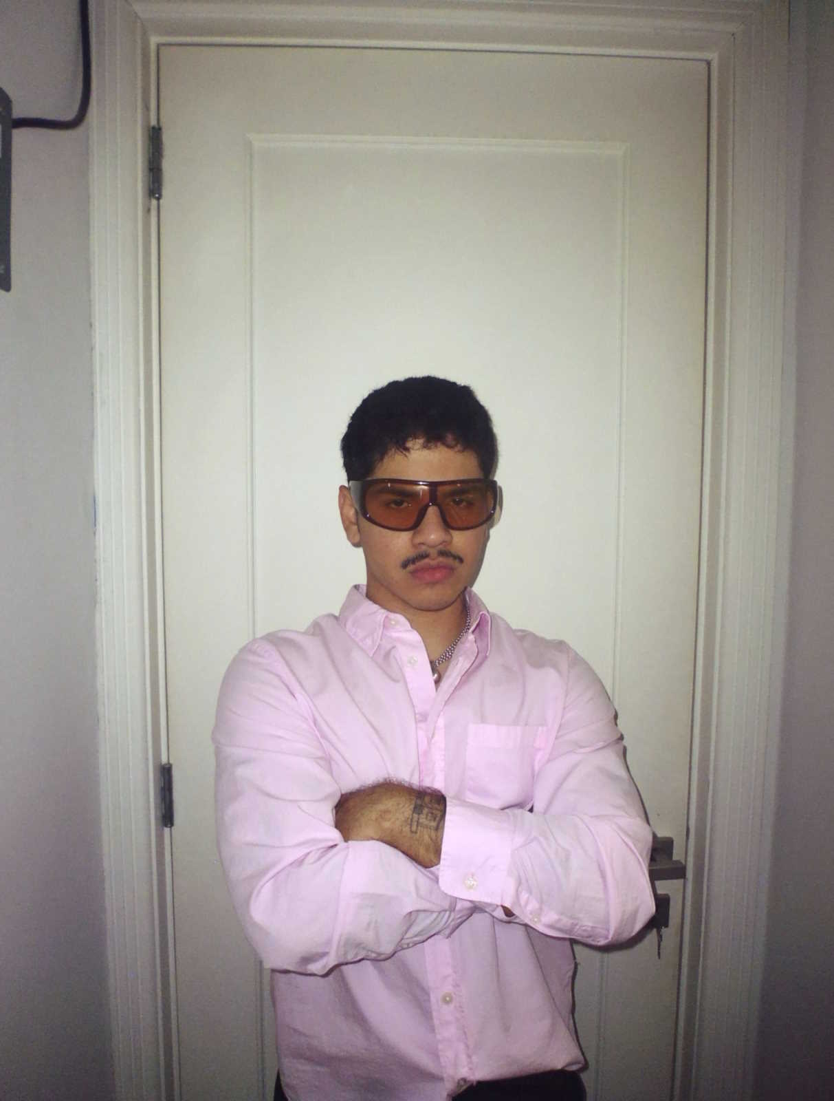 Promotional image of Andry Kiddos. He is standing in front of a white door. He is wearing a pink button-down shirt and large amber sunglasses.