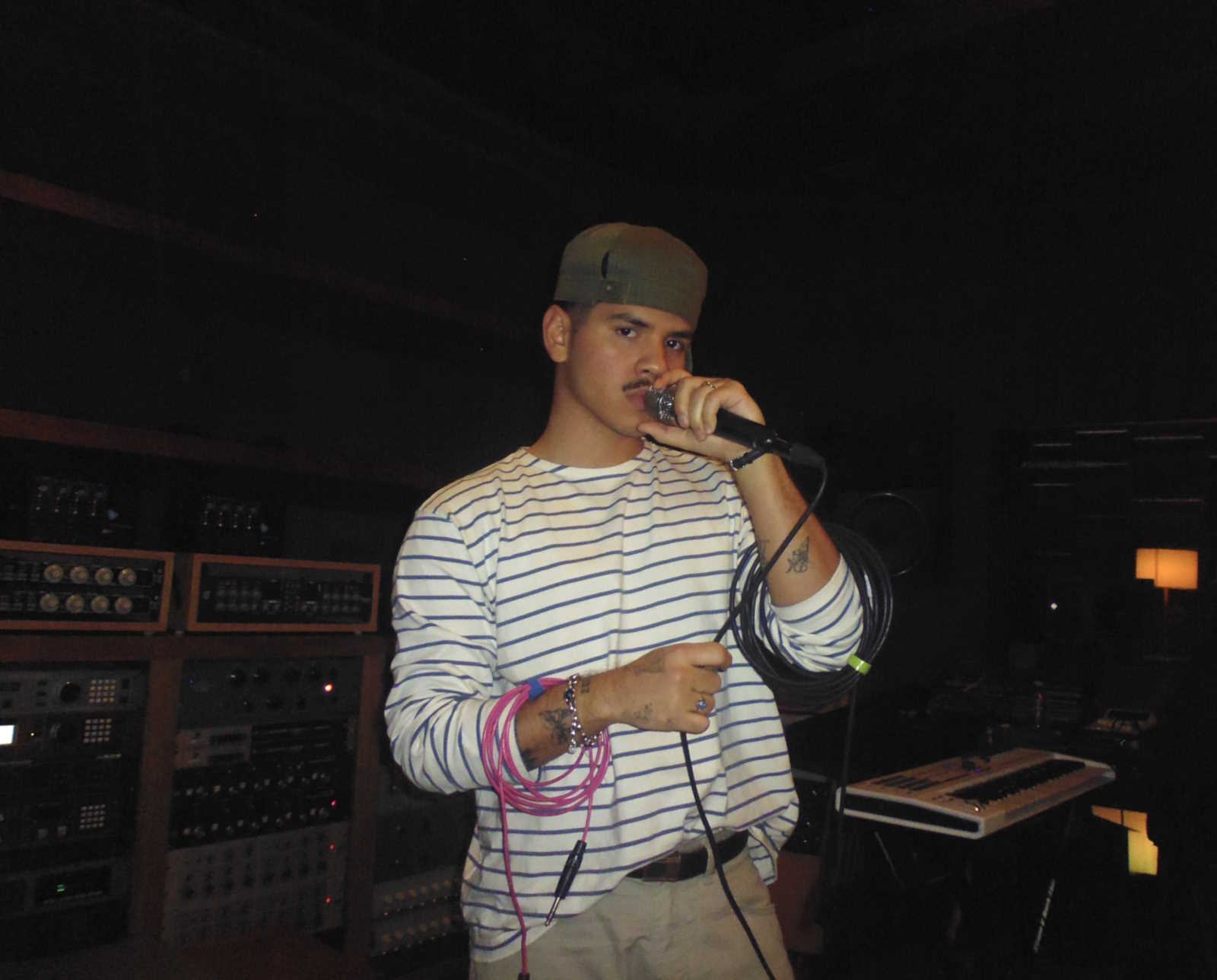 Promotional image of Andry Kiddos. He is standing. The background is dark. He is holding a microphone. He is wearing an olive green cap and wearing a long-sleeve white shirt with dark horizontal stripes.