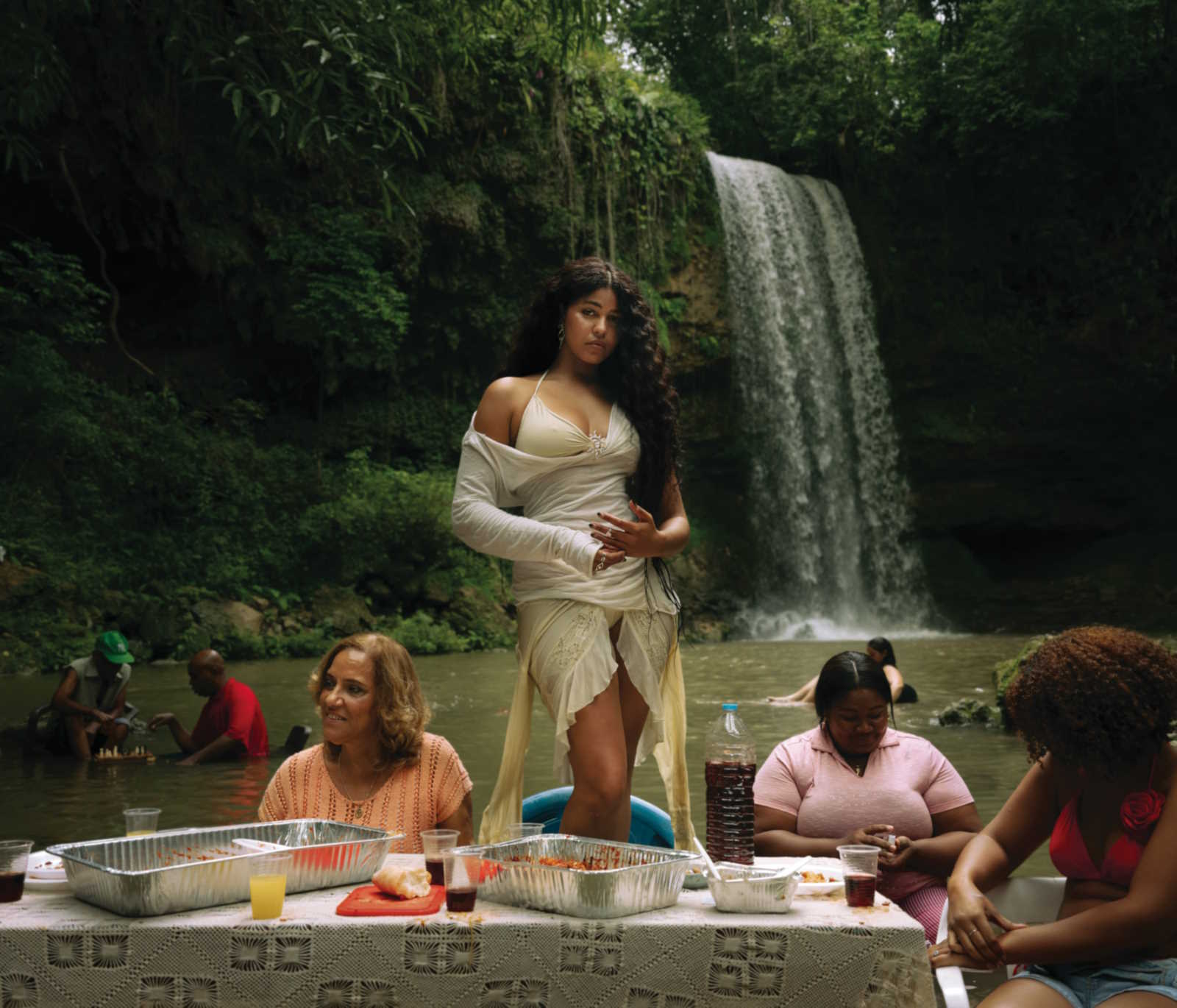 A promotional image of Yendy standing on the shore of a lake. The lake is in the background, and a waterfall is visible in the distant background. Multiple other people are in the image, and a table with food appears in the foreground.