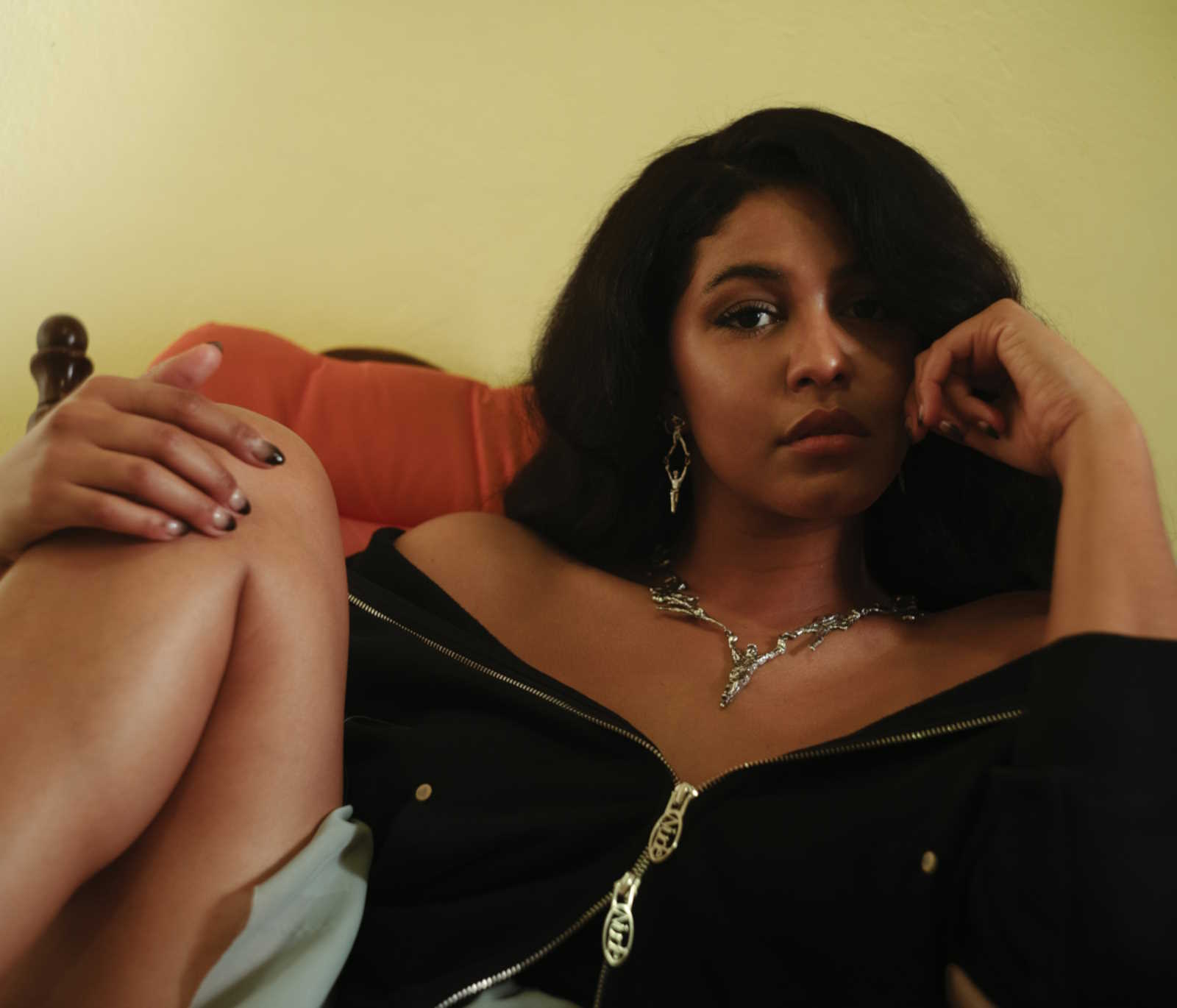 A promotional image of YEИDRY seated in a soft chair or sofa. She is in a relaxed pose, with one knee raised and her hand on her knee. She is wearing a black shirt.